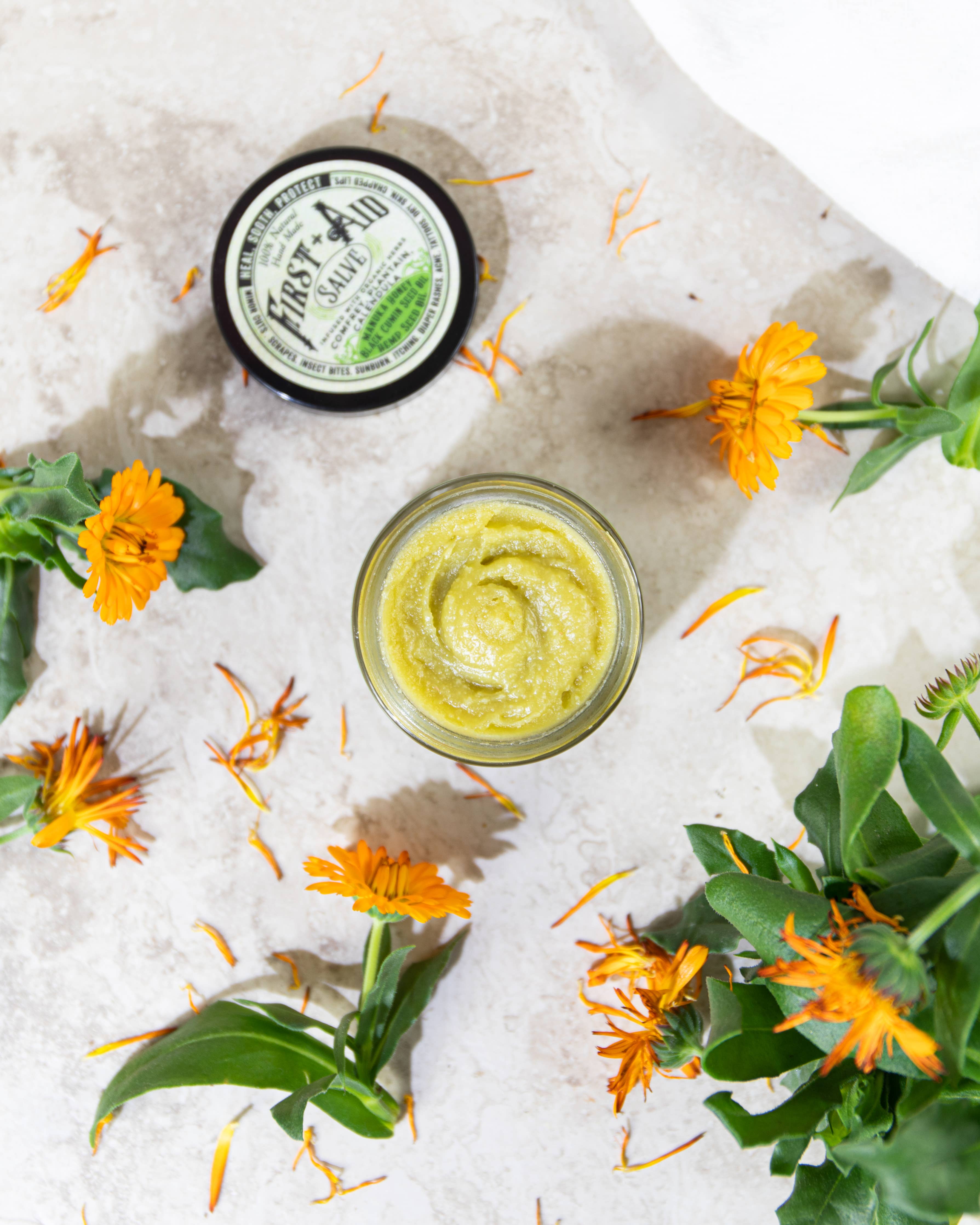 An open jar of Roots And Leaves First Aid Salve rests on a light surface, surrounded by vibrant calendula flowers and petals, with the product’s labeled lid nearby.