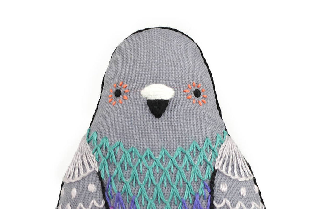 A Kiriki Press Pigeon - Embroidery Kit soft toy with a gray head, orange markings near the eyes, and intricate turquoise and purple stitching on its chest.