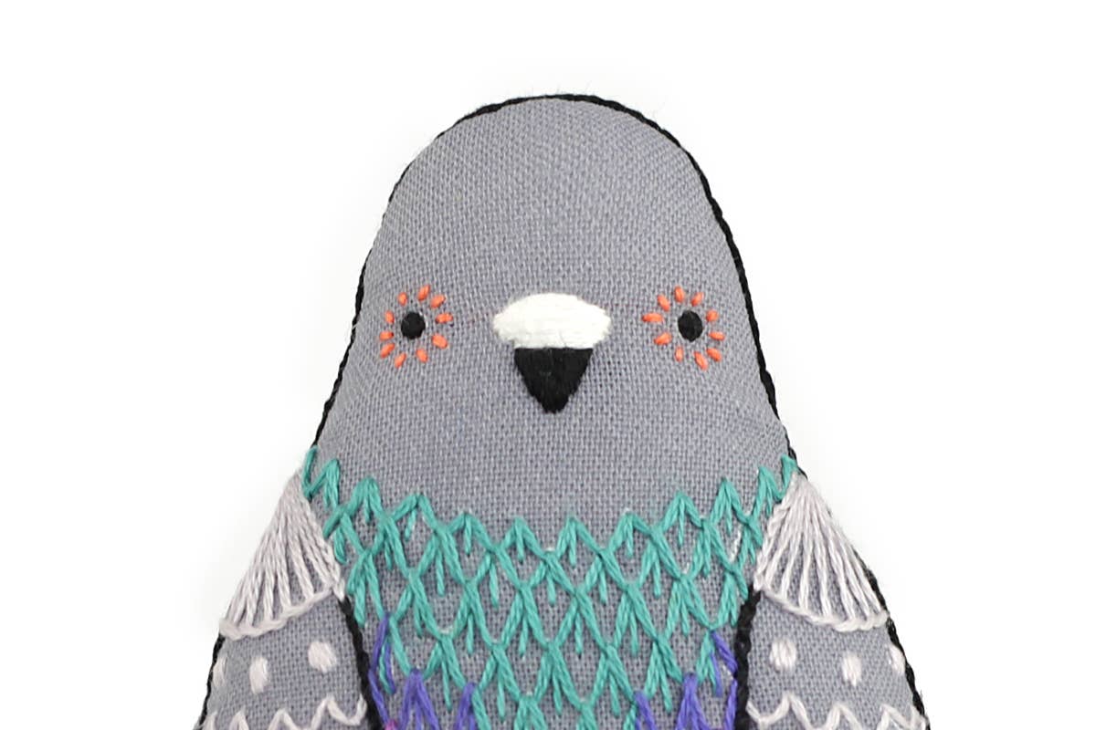 A Kiriki Press Pigeon - Embroidery Kit soft toy with a gray head, orange markings near the eyes, and intricate turquoise and purple stitching on its chest.
