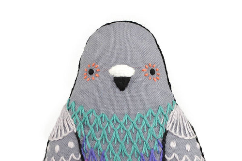 A Kiriki Press Pigeon - Embroidery Kit soft toy with a gray head, orange markings near the eyes, and intricate turquoise and purple stitching on its chest.