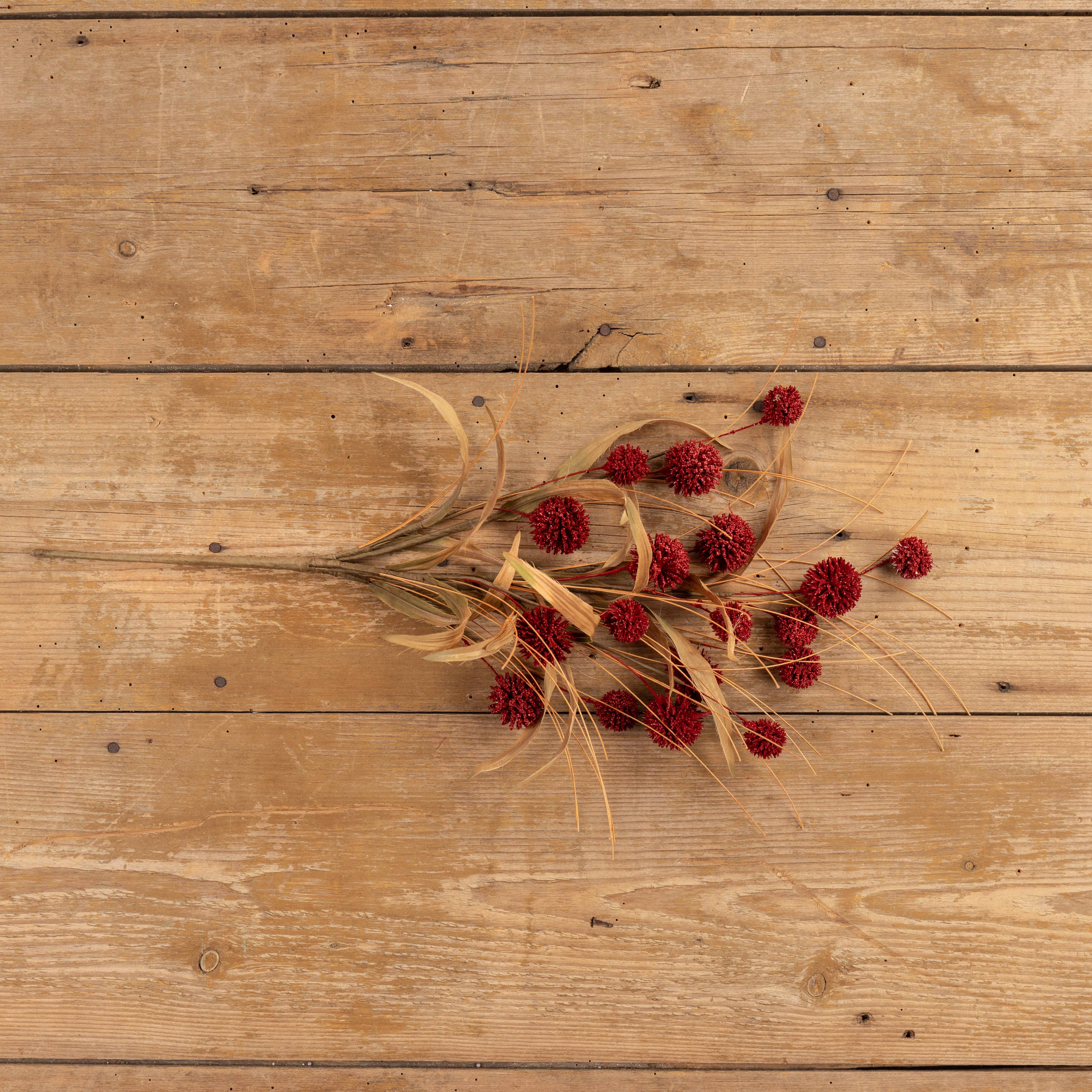 A 23" BURGUNDY POM POM BUSH by Ragon House, paired with dried red flowers and beige grasses, is arranged on a rustic wooden surface with visible grain and knots.