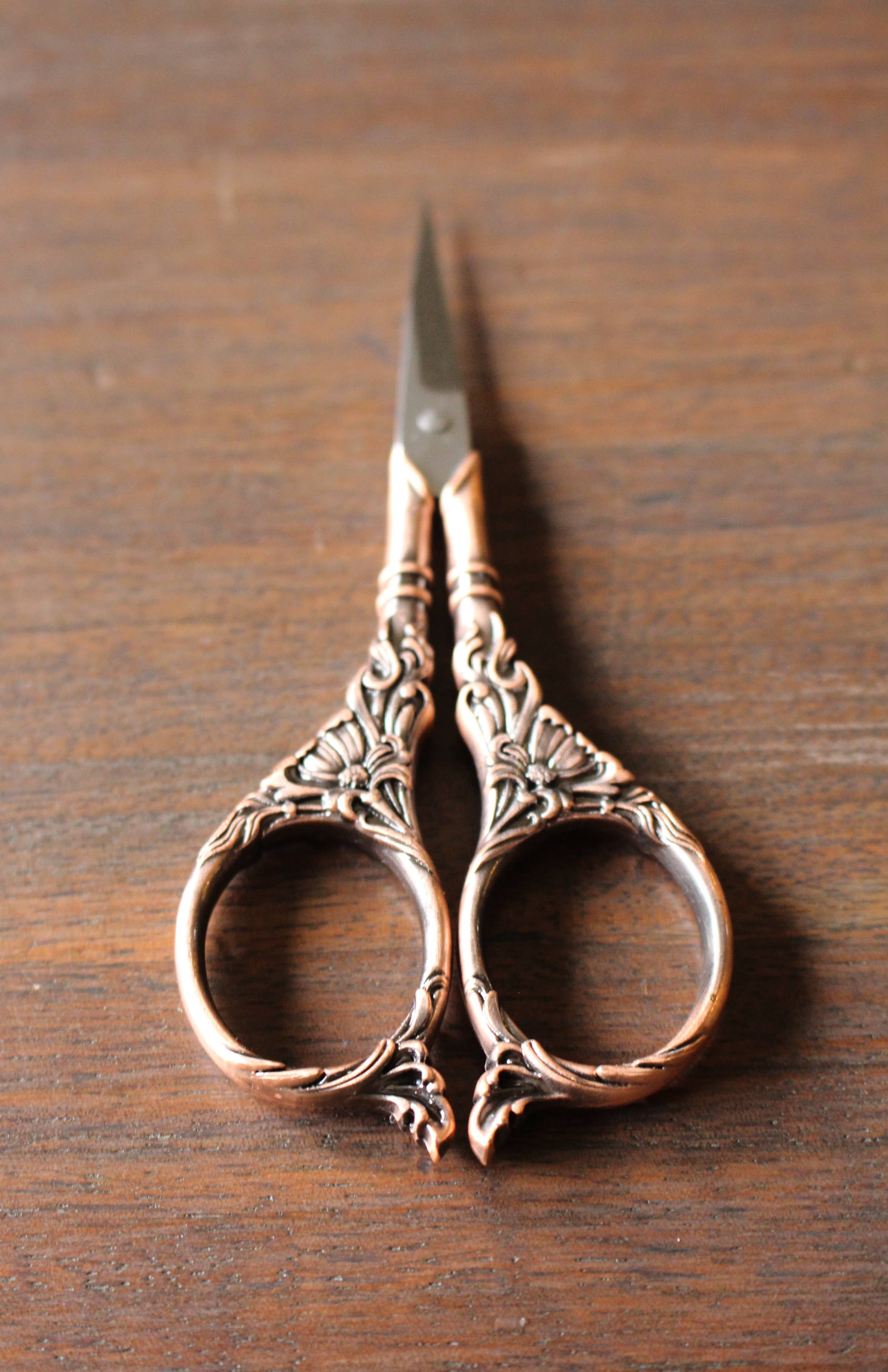 The NNK Press Botanical Garden Scissors Antique Gold, featuring botanical handles and intricate vintage-style metalwork, rest on a wooden surface.