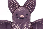 A close-up of the Bat - Embroidery Kit by Kiriki Press, a DIY plushie made of purple fabric with stitched pink ear and wing details, a smiling face with white fangs, and a pink nose.