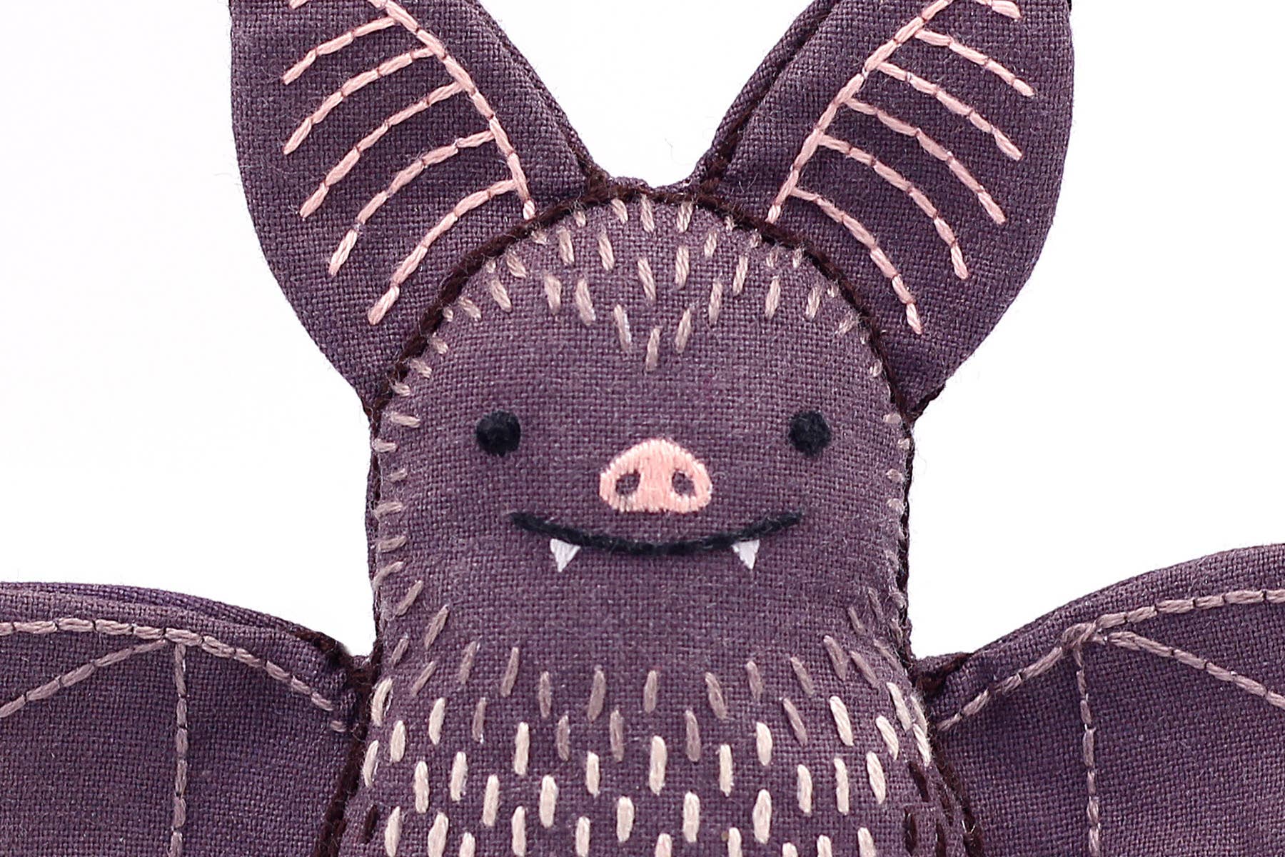 A close-up of the Bat - Embroidery Kit by Kiriki Press, a DIY plushie made of purple fabric with stitched pink ear and wing details, a smiling face with white fangs, and a pink nose.