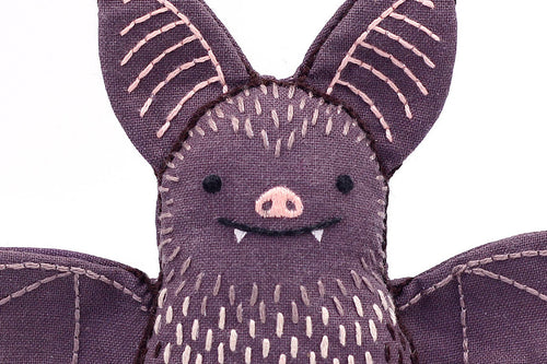 A close-up of the Bat - Embroidery Kit by Kiriki Press, a DIY plushie made of purple fabric with stitched pink ear and wing details, a smiling face with white fangs, and a pink nose.