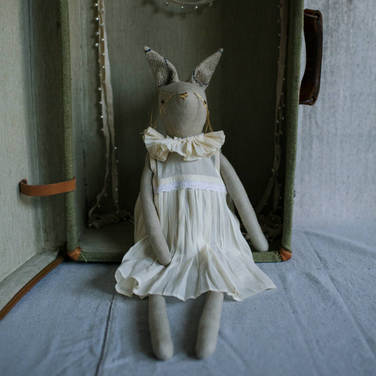 The Skippy Cotton Ruffle Bun Ragdoll—an art doll with hand-stitched details, gray ears, and a cotton dress—sits inside an open vintage suitcase lined with fabric.