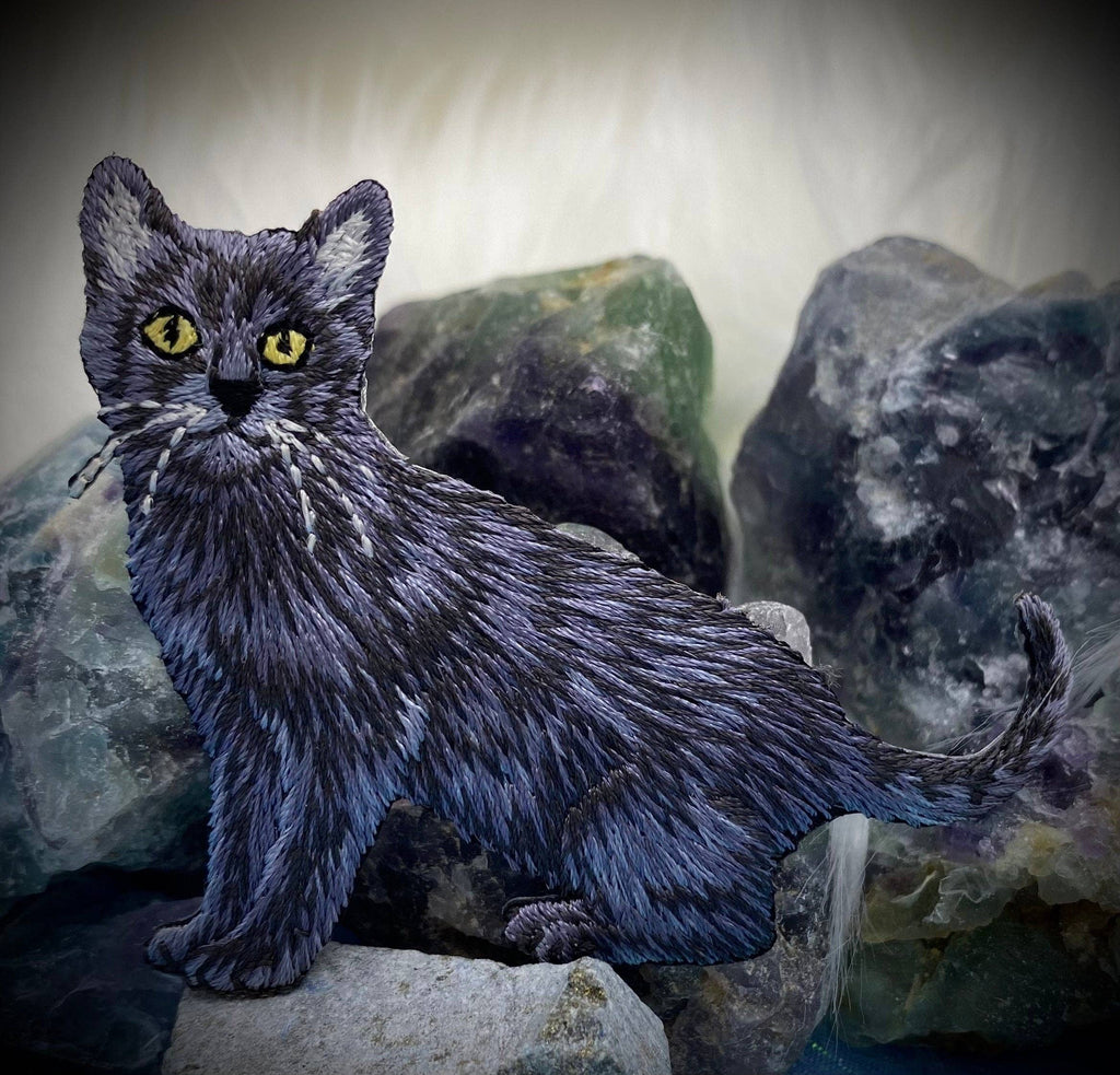 The Black Cat Realistic Full Body Iron on Patch by Wholesale Applique features detailed embroidery and vivid yellow eyes, standing out against a blurred backdrop of colorful stones for a striking pet-inspired design.