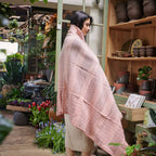 Wrapped in the Silica Rose Throw in shades of salmon gray stripes from Variously, a person stands cozily amid a plant-filled shop, surrounded by flowers, potted plants, and wooden shelves displaying pottery and books.