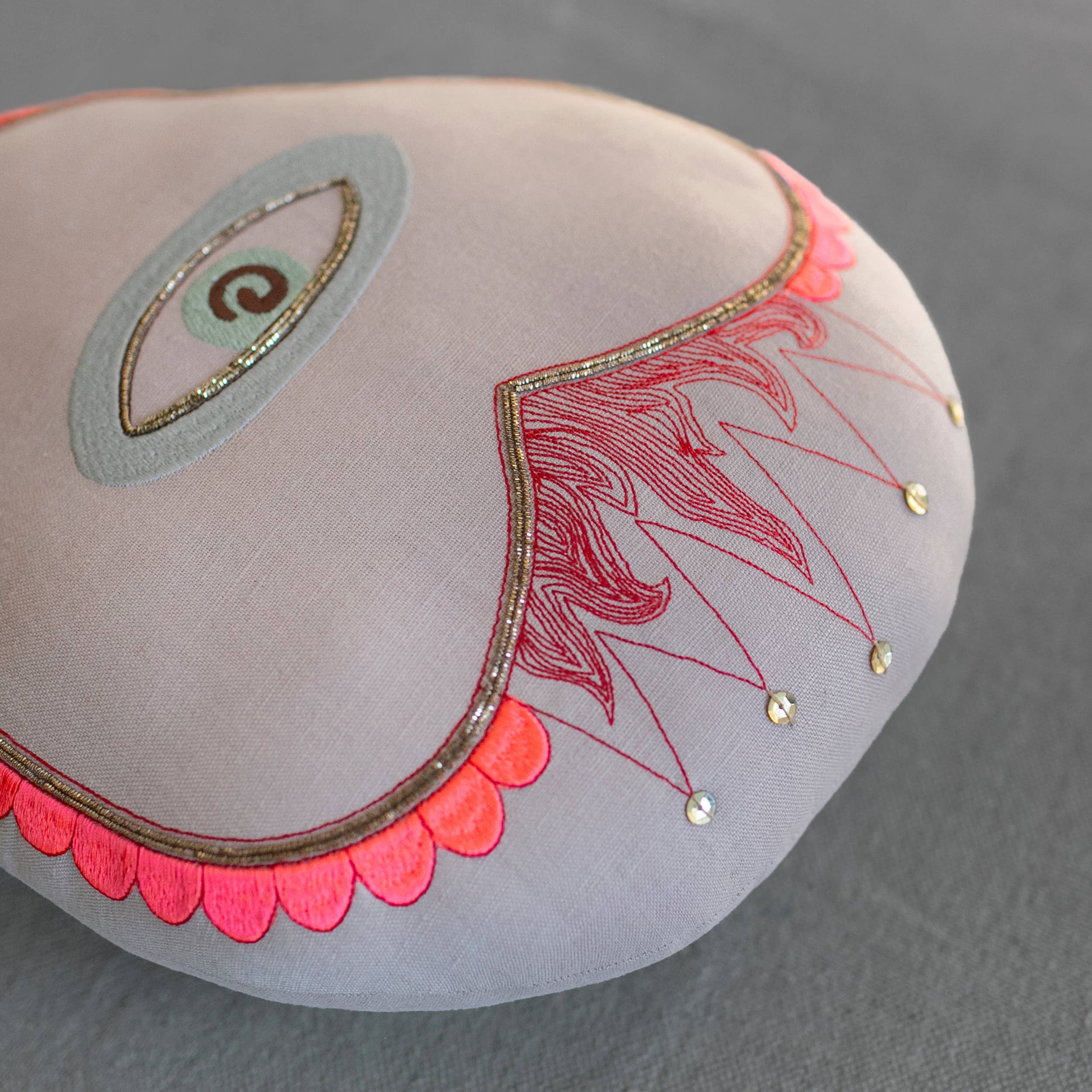 The Skippy Cotton Sacred Heart Pillow is a handmade, cotton-filled round cushion in light gray linen, featuring abstract eye and heart embroidery with red and pink details—perfect for adding whimsy to your decor.