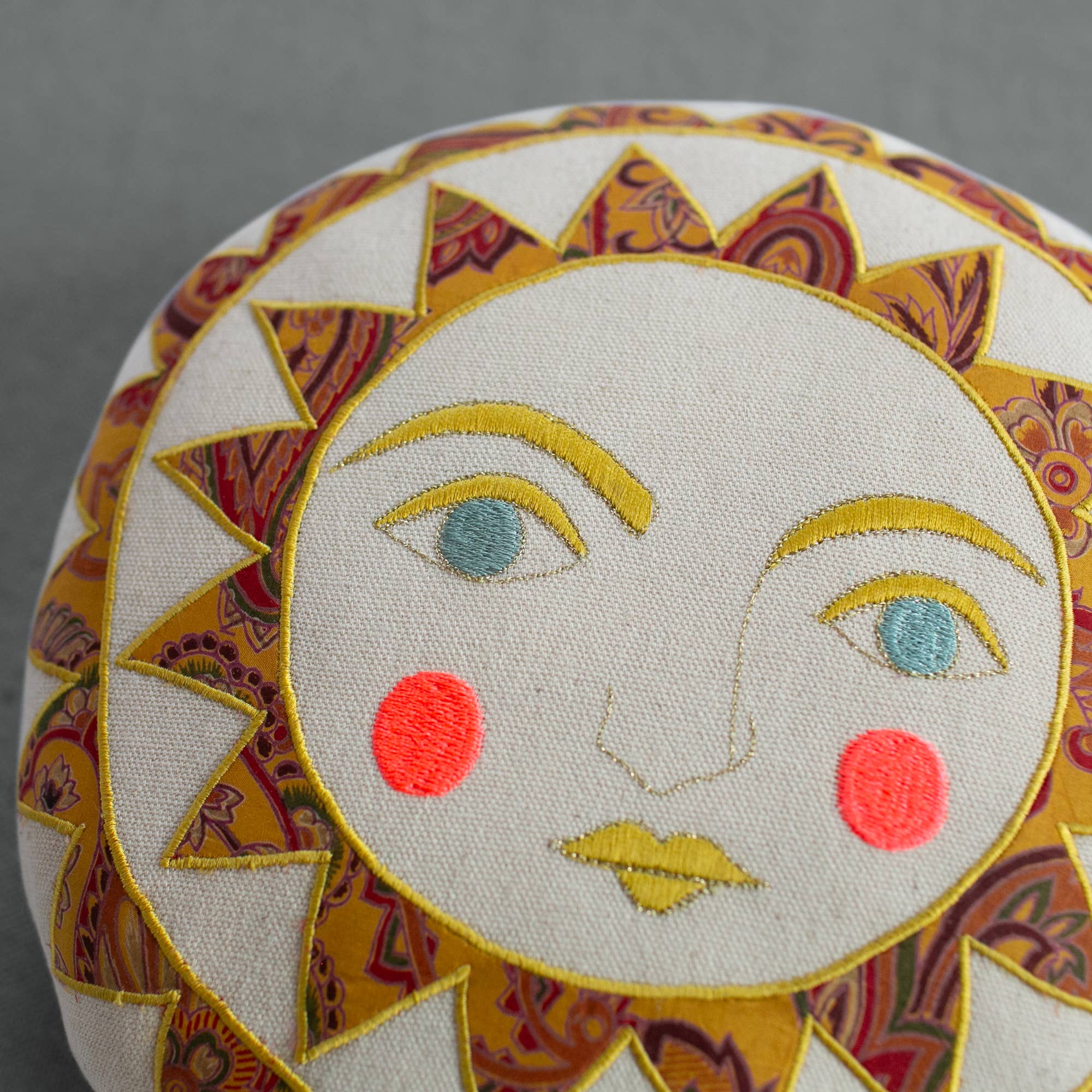 The Skippy Cotton Bright Neon Sun Pillow is a handmade, cotton-filled round cushion featuring an embroidered sun face with yellow and orange rays, blue eyes, yellow lips, and red cheeks—a vibrant accent for whimsical decor.