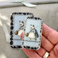 A small hand holds a Firefly Notes Kitty notions tin: Small, featuring Business Cat in a dress with a basket and a blue and white floral border. The tin clicks shut, while another similar tin is blurred in the background.
