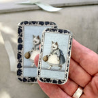 A small hand holds a Firefly Notes Kitty notions tin: Small, featuring Business Cat in a dress with a basket and a blue and white floral border. The tin clicks shut, while another similar tin is blurred in the background.