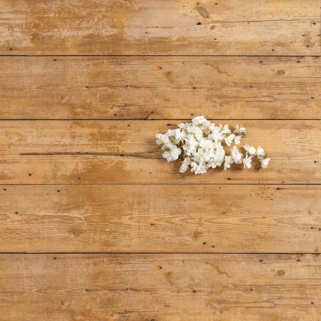 A FAUX WHITE CHERRY BLOSSOM SPRAY from Ragon House rests horizontally on a rustic wooden surface with visible grain and knots.