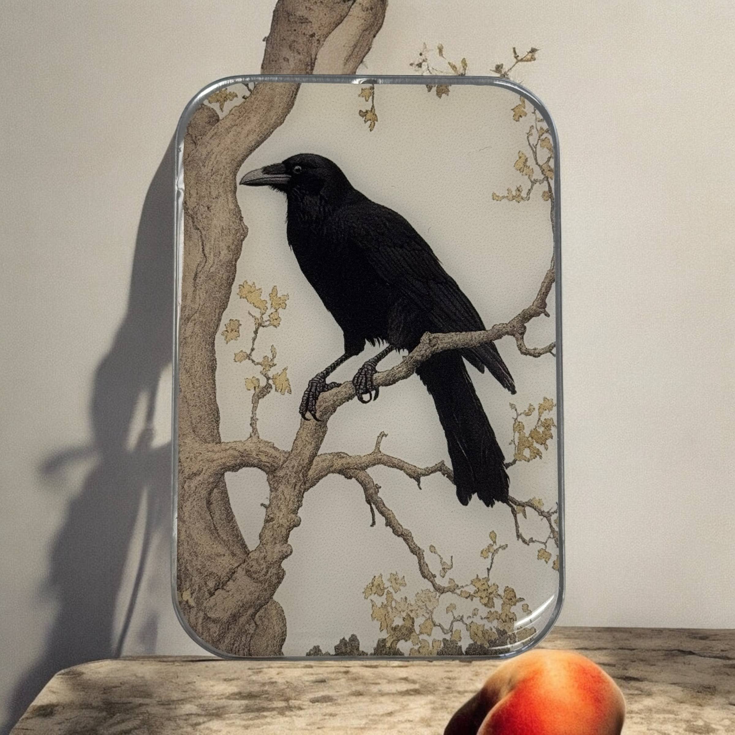 A handcrafted Firefly Notes Black Crow Notions Tin sits in front of a framed black crow illustration and a peach, with the peach casting a partial shadow on the surface.