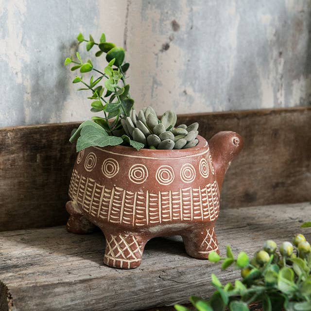 The Vagabond Vintage CEMENT RED TURTLE PLANTER (Large), shaped like a turtle with geometric patterns, holds a succulent and leafy plant on a rustic wooden surface against a weathered wall.