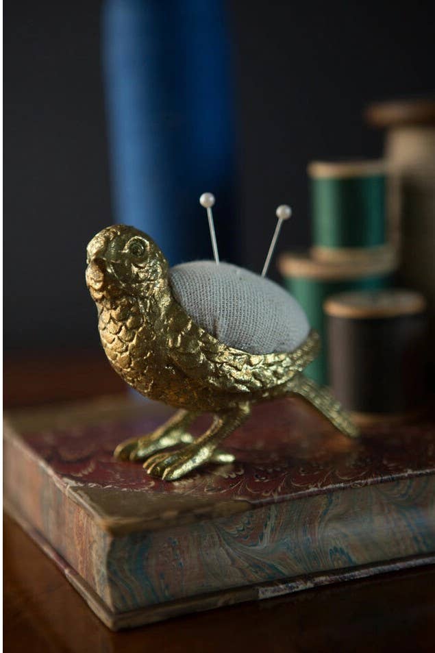 A gold Bird Needle Holder by Vagabond Vintage, with two pins in its back, sits atop a decorative book, while spools of thread are softly blurred in the background.