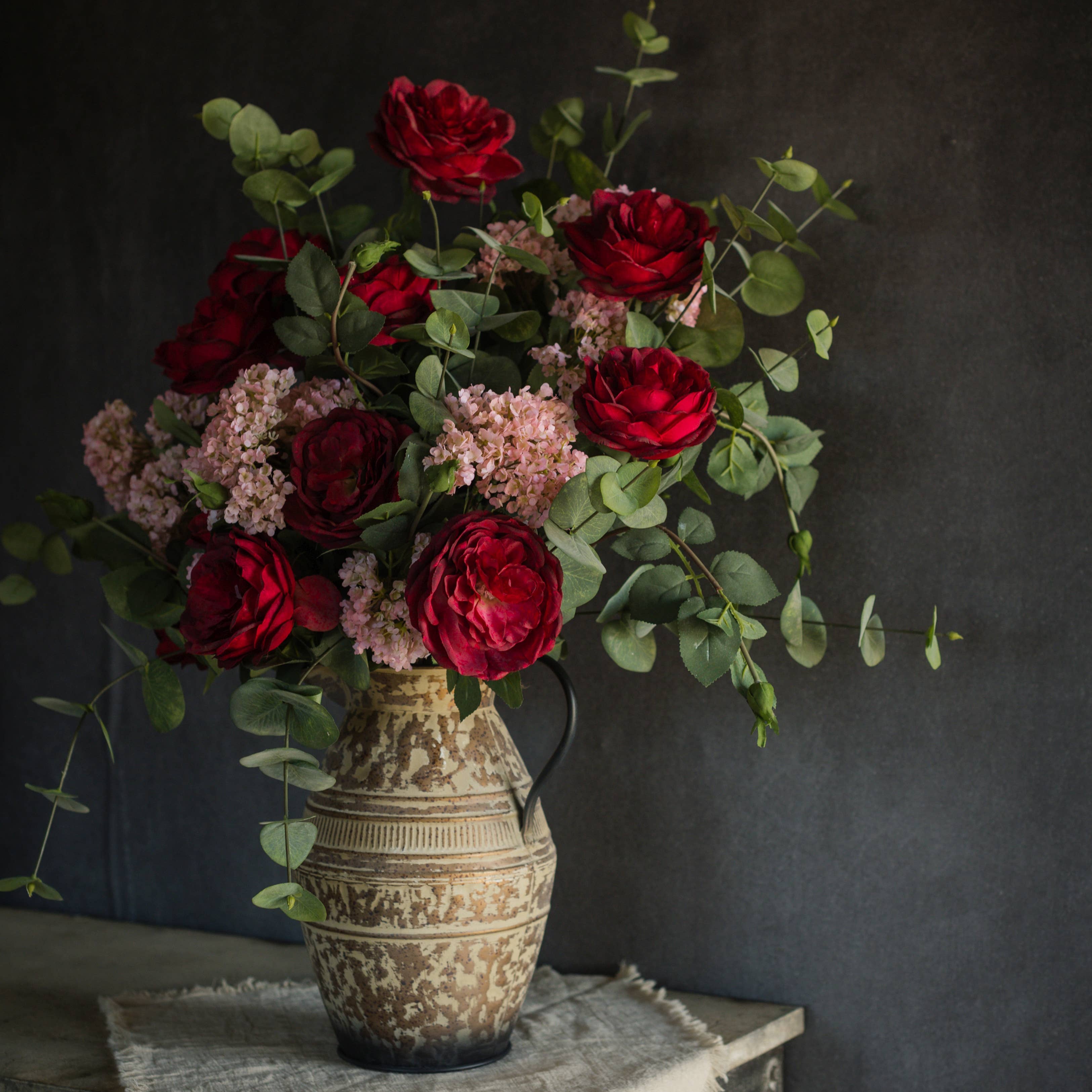A rustic ceramic jug holds red roses, pink blooms, green eucalyptus, and a 25" PINK FAUX HYDRANGEA SPRAY from Ragon House, all arranged on textured cloth against a dark neutral background.