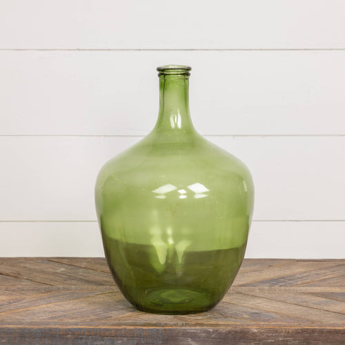 The Ragon House 16" GREEN GLASS BOTTLE, featuring a narrow neck and rounded body, makes a striking centerpiece on a wooden surface against a white shiplap wall.