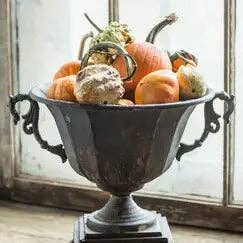 The Vagabond Vintage Distressed Black Iron Urn, filled with assorted small pumpkins and gourds, sits on a windowsill bathed in natural light.