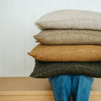 A person in blue jeans sits on a wooden bench, their upper body and face hidden behind a stack of three large Indaba Trading 24x24 Lina Linen Pillow Avocado: Cover with Filler pillows in beige, brown, and dark gray.