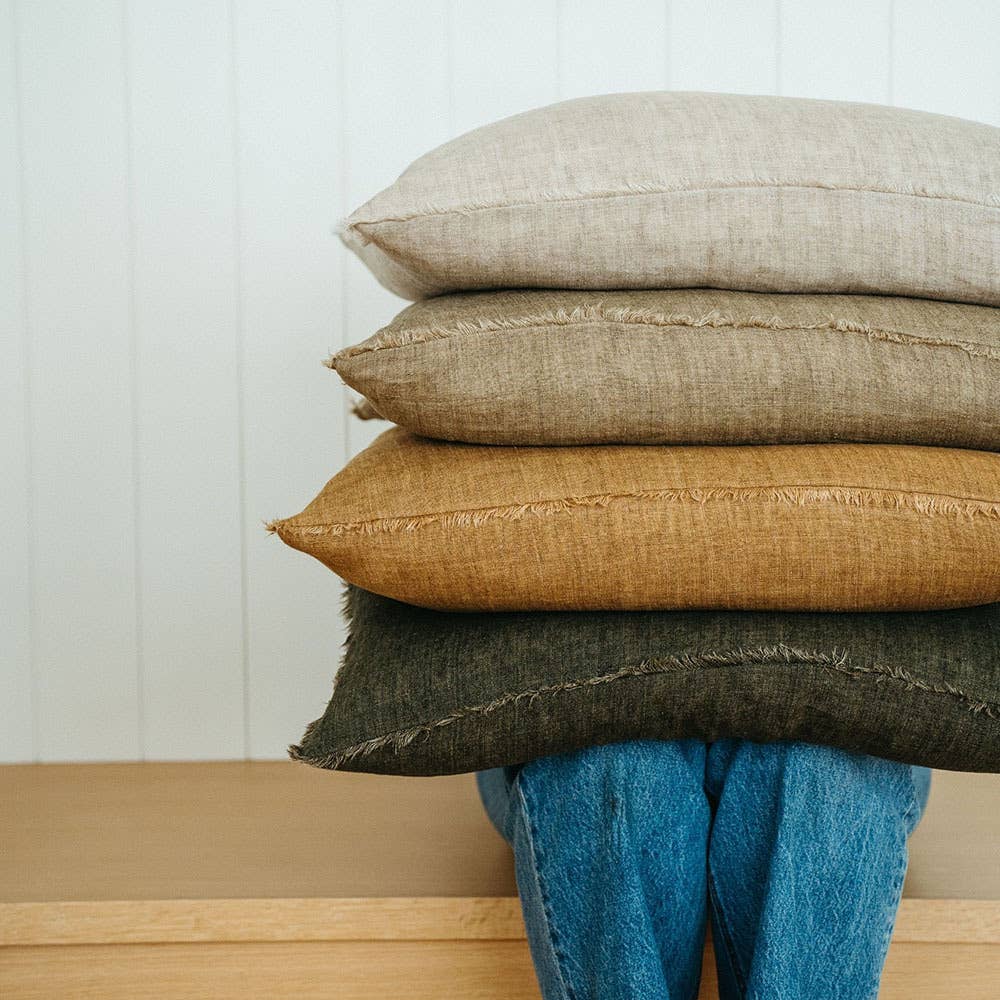 A person in blue jeans sits on a wooden bench, their upper body and face hidden behind a stack of three large Indaba Trading 24x24 Lina Linen Pillow Avocado: Cover with Filler pillows in beige, brown, and dark gray.