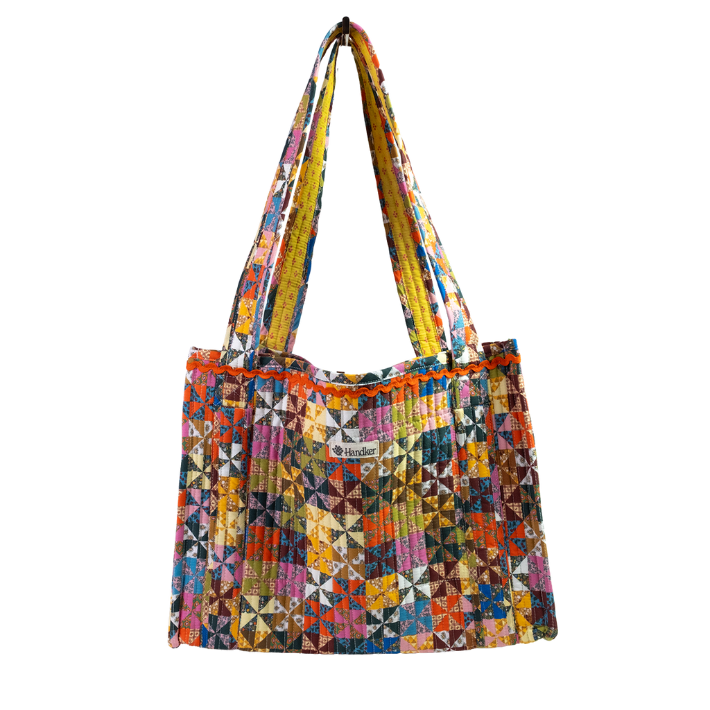 The Calico Quilted Tote by Handker Bandanas features a geometric pinwheel quilt pattern in bright colors, two shoulder straps, and a small kinder label on the front against a plain white background.