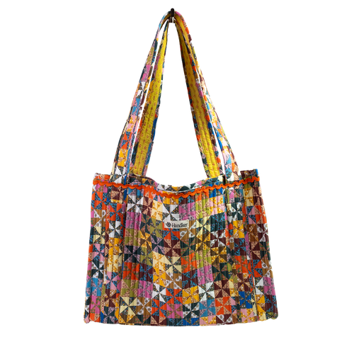 The Calico Quilted Tote by Handker Bandanas features a geometric pinwheel quilt pattern in bright colors, two shoulder straps, and a small kinder label on the front against a plain white background.