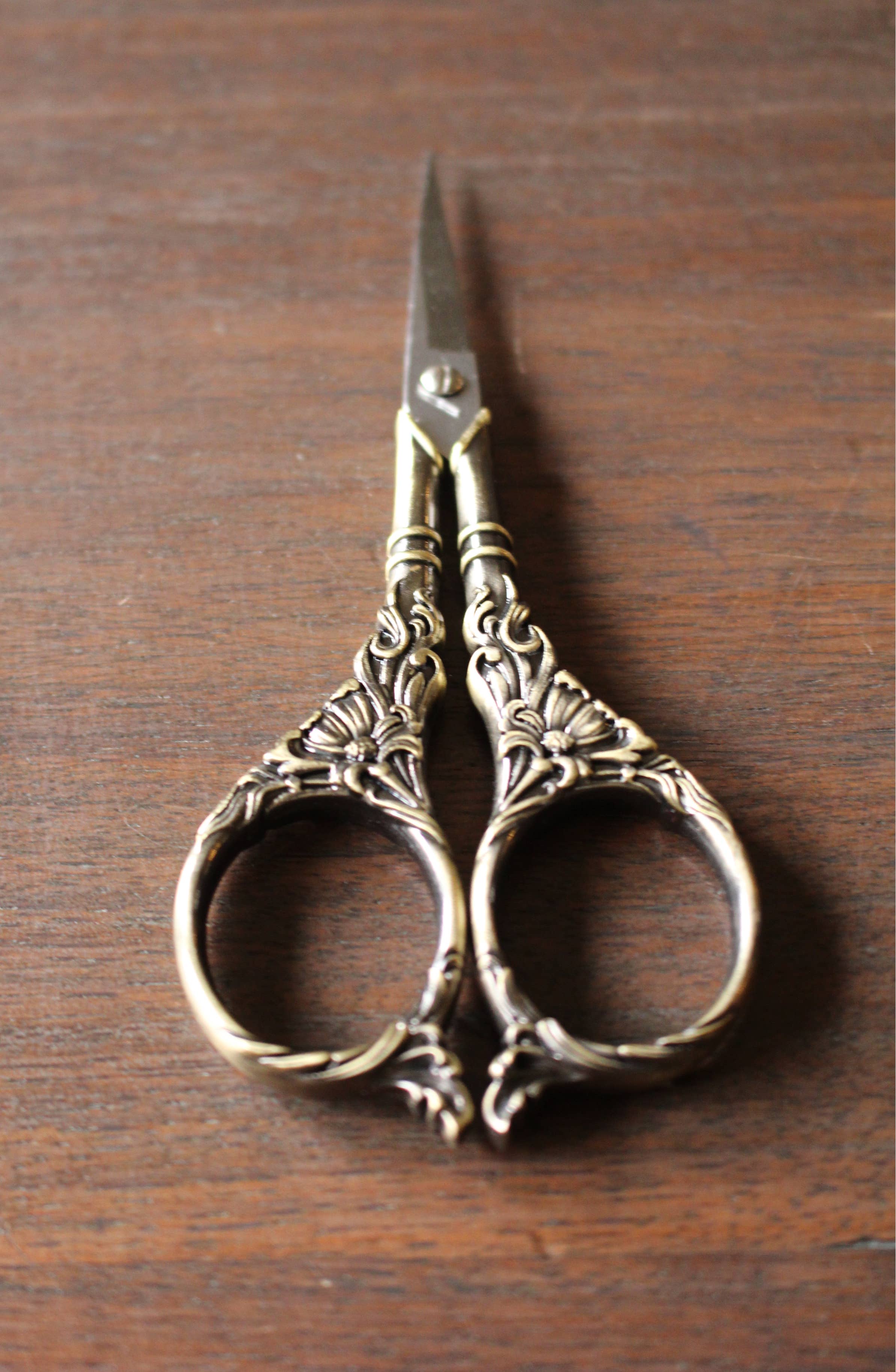 The NNK Press Botanical Garden Scissors Antique Gold, featuring intricate botanical handles and stainless steel blades, rests on a wooden surface.