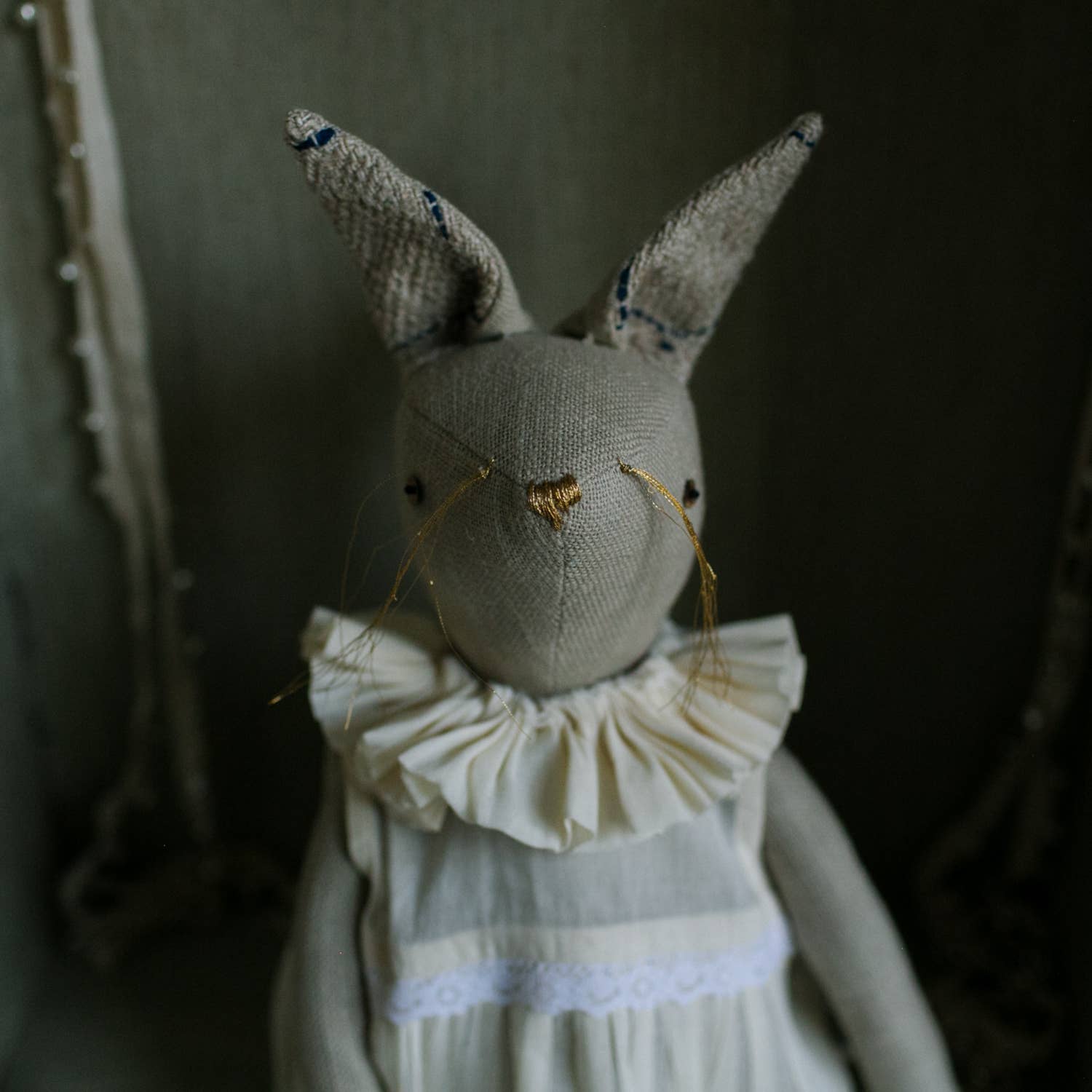 The Skippy Cotton Ruffle Bun Ragdoll is a handmade art doll featuring a cotton-filled body, long ears, a ruffled white collar, simple hand-stitched details, and a cotton dress, shown against a dim background.