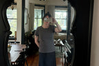A person with dark hair wears a Thyne Goods Brown Ojai Tee and a black skirt, taking a mirror selfie in a cozy room with wooden floors, a round table, chairs, and a sofa by the window with curtains and plants.