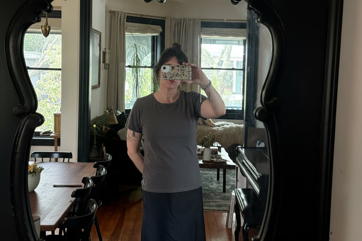 A person with dark hair wears a Thyne Goods Brown Ojai Tee and a black skirt, taking a mirror selfie in a cozy room with wooden floors, a round table, chairs, and a sofa by the window with curtains and plants.