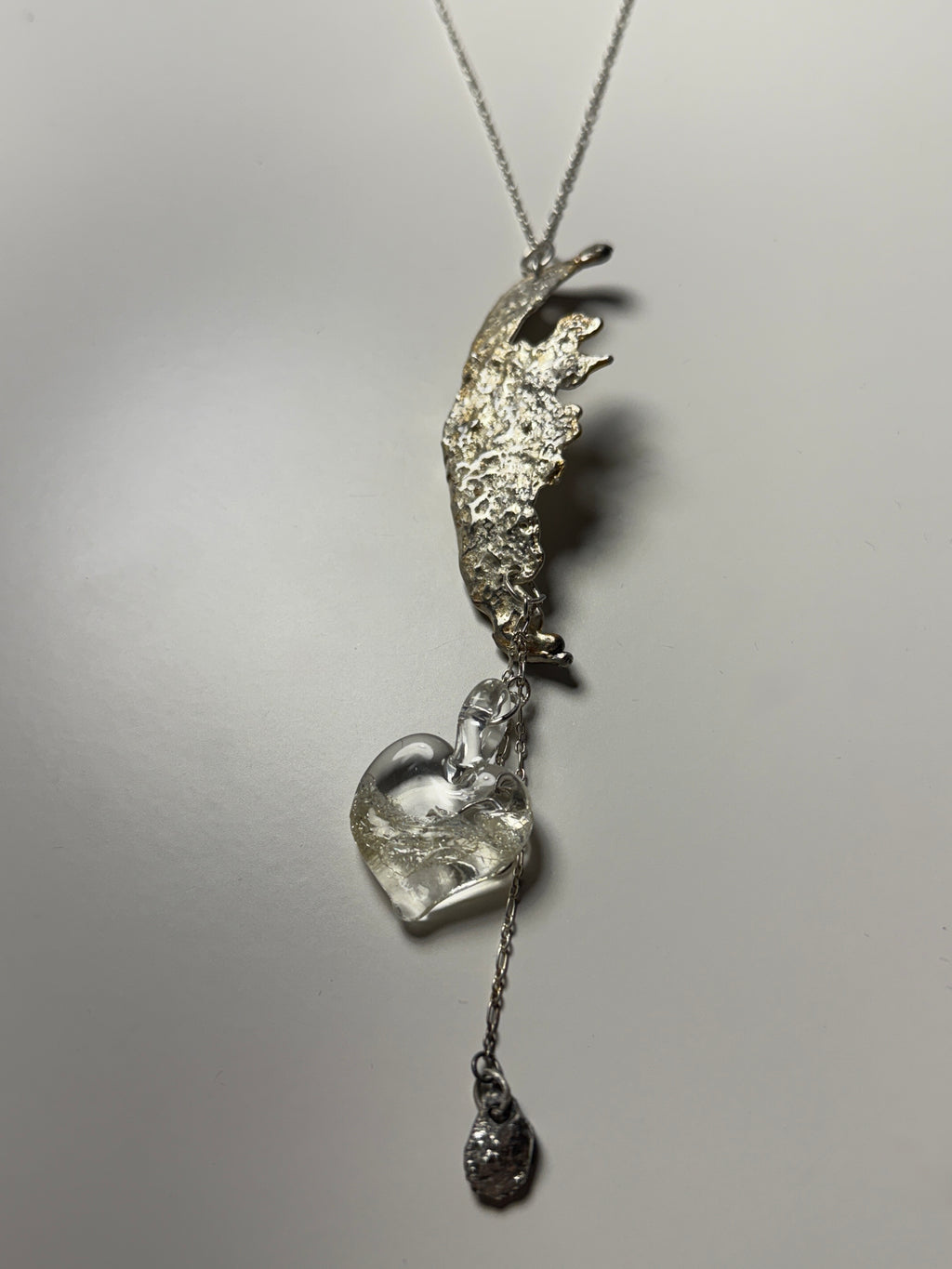 A silver necklace from Thyne Goods, designed by Ash Espinoza x Thyne Studio, with an abstract textured metal look, a handmade glass heart pendant, and a dangling charm on a delicate chain. Shown against a plain white background.