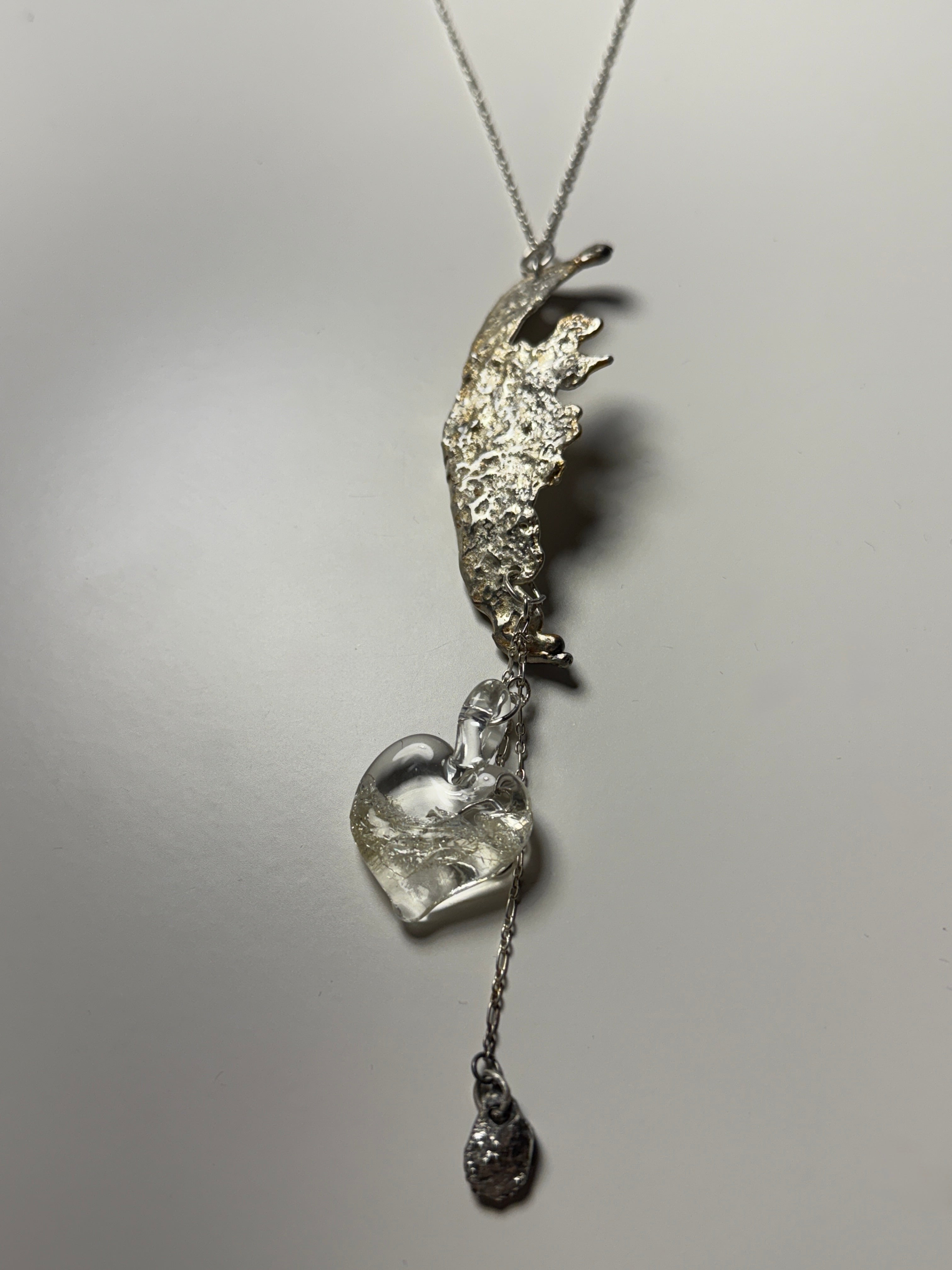 A silver necklace from Thyne Goods, designed by Ash Espinoza x Thyne Studio, with an abstract textured metal look, a handmade glass heart pendant, and a dangling charm on a delicate chain. Shown against a plain white background.