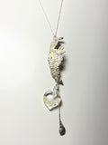 The Wing Fall necklace by Thyne Goods (Ash Espinoza x Thyne Studio) features an abstract textured pendant, a handmade glass heart with silver, and a small dangling charm, all shown on a white background.