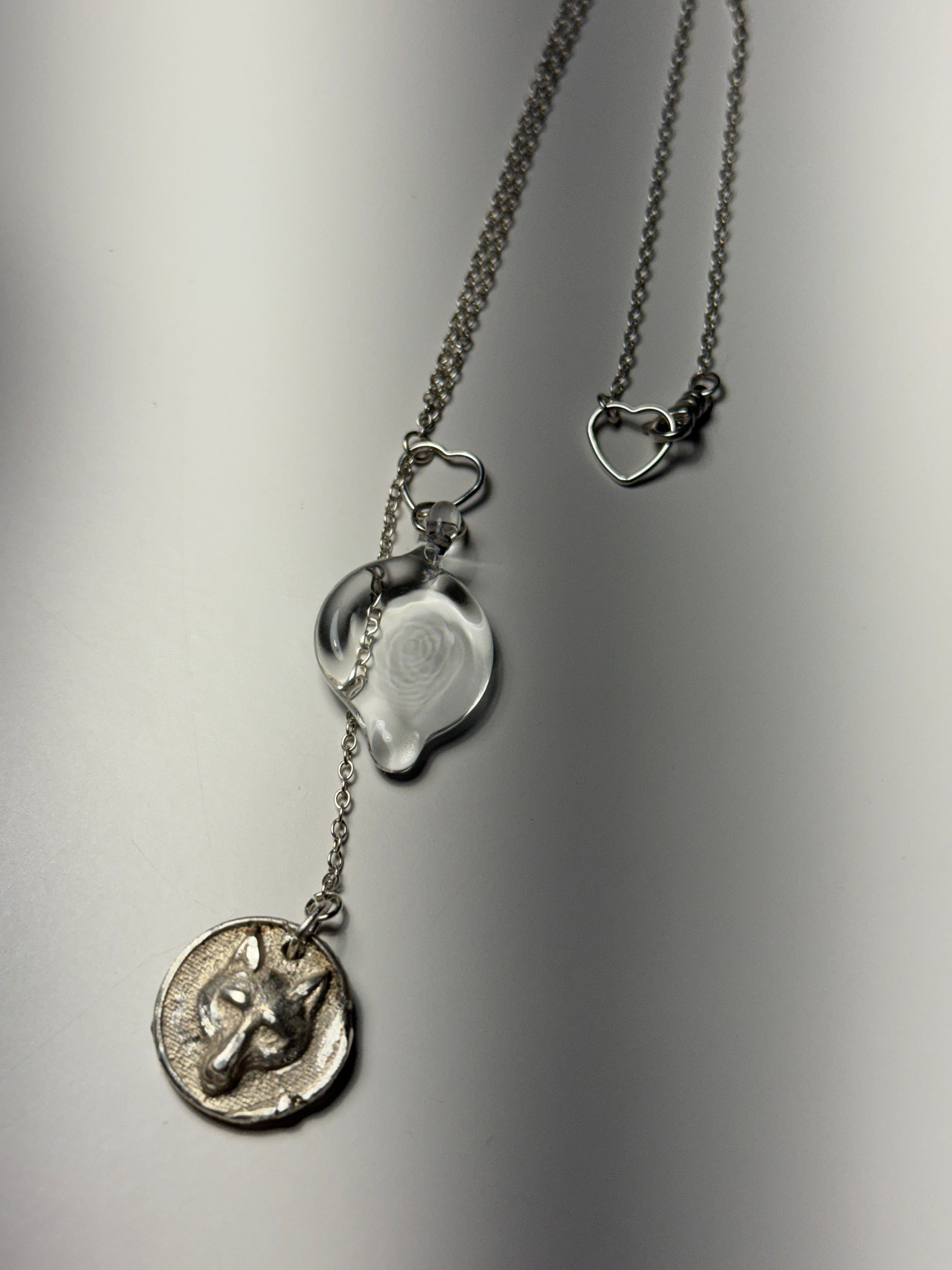 The Trickster • Ash Espinoza x Thyne Studio necklace by Thyne Goods features a sterling silver chain with a heart-shaped clasp, a clear handmade glass heart pendant, and a round wolf head charm, displayed on a smooth gray surface.